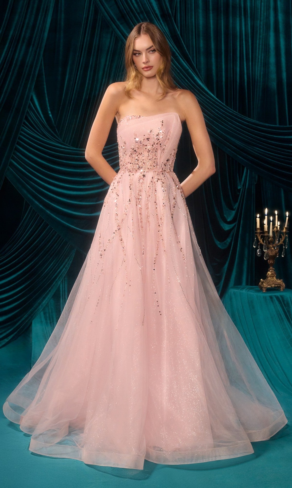 A woman stands before teal draped curtains wearing the pink Long Prom Dress: Andrea and Leo A1491, featuring sequin embellishments and a sheer tulle overlay. A candelabra appears on the right.